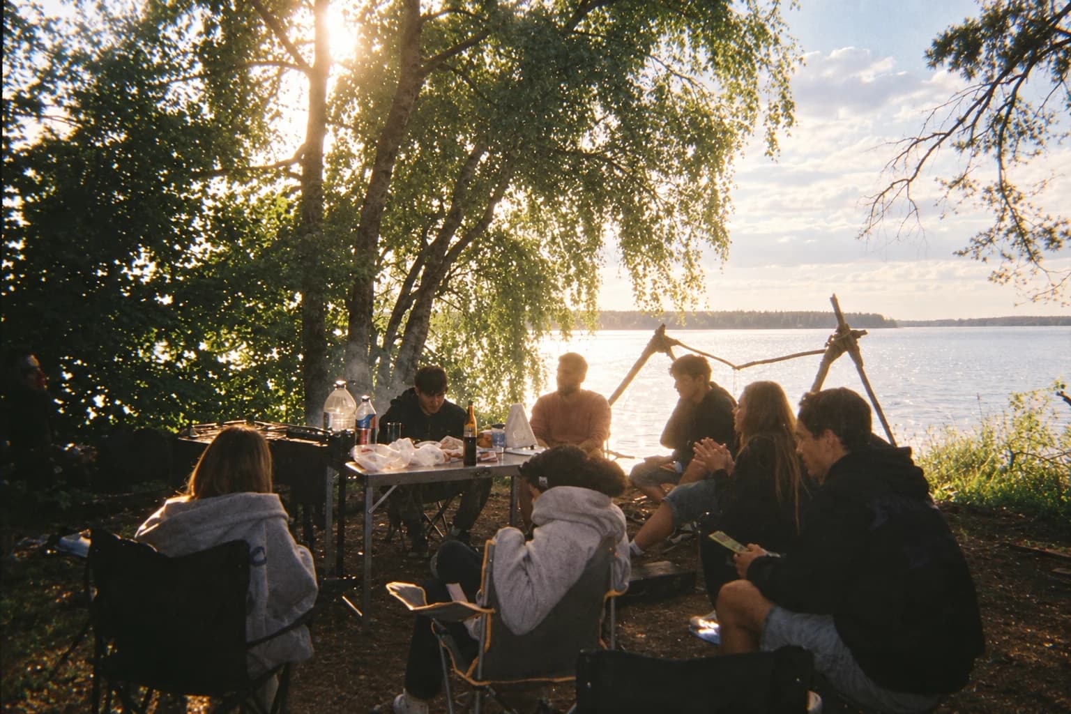 Friends gathered around a picnic table beneath sunlit trees beside a lake